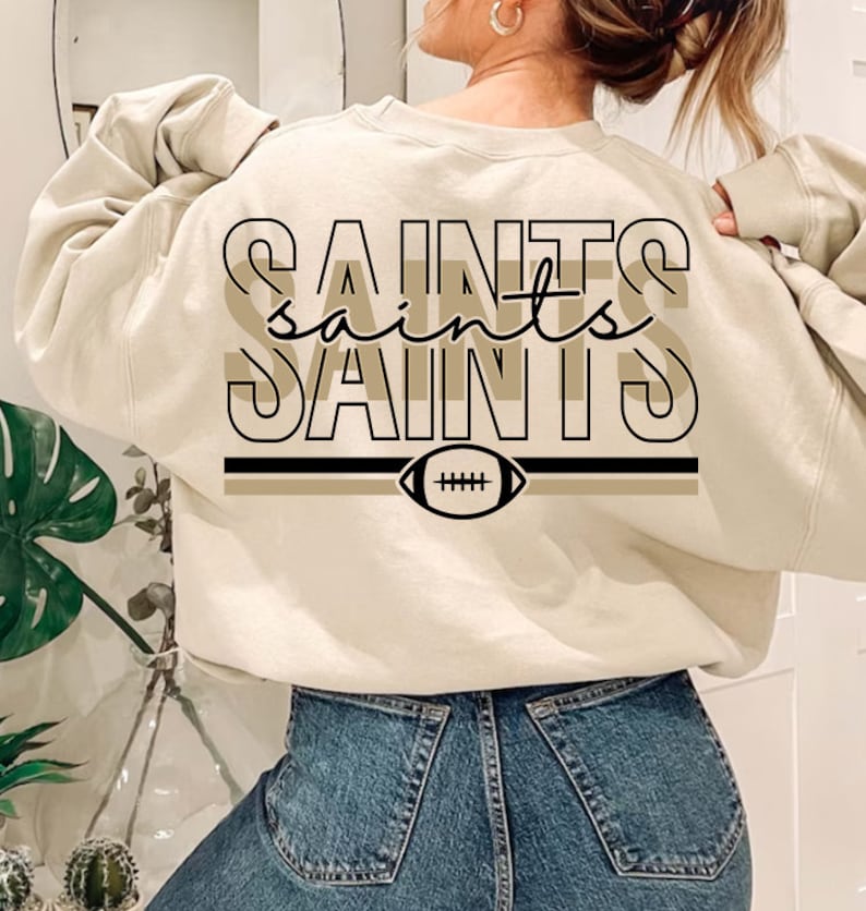 May include: A beige sweatshirt with a black and brown graphic design that says "Saints" with a football graphic underneath.