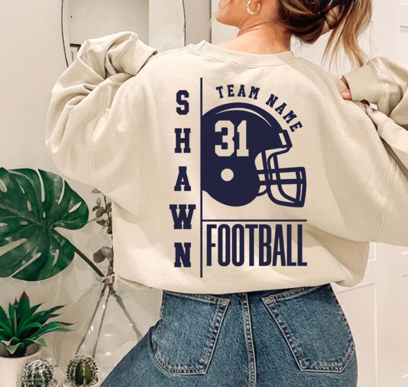 May include: A cream-colored sweatshirt with a navy blue graphic on the back. The graphic features a football helmet with the number 31, the words "TEAM NAME" and "FOOTBALL", and the letters "SHAWN" vertically down the side.