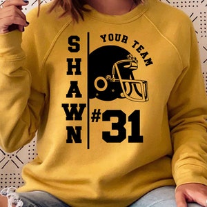 K&ouml;nnte beinhalten: Ein senfgelber Pullover mit schwarzem Text, der "SHAWN YOUR TEAM #31" lautet, und einer schwarzen Grafik eines Footballhelms.