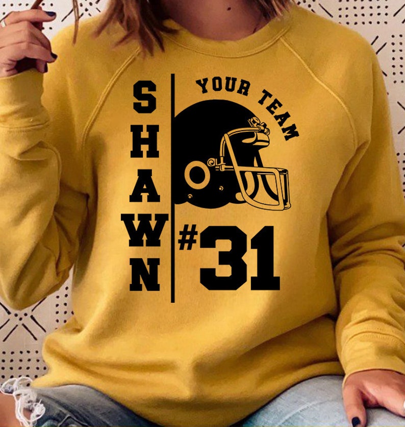 K&ouml;nnte beinhalten: Ein senfgelber Pullover mit schwarzem Text, der "SHAWN YOUR TEAM #31" lautet, und einer schwarzen Grafik eines Footballhelms.