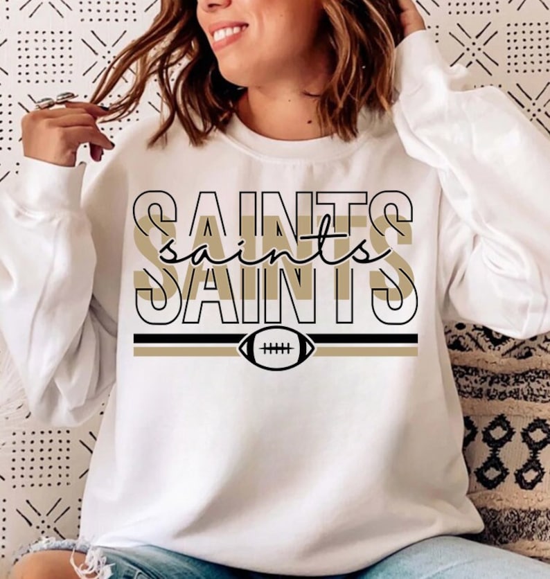 May include: White sweatshirt with a black and gold graphic that says "Saints" with a football graphic underneath.
