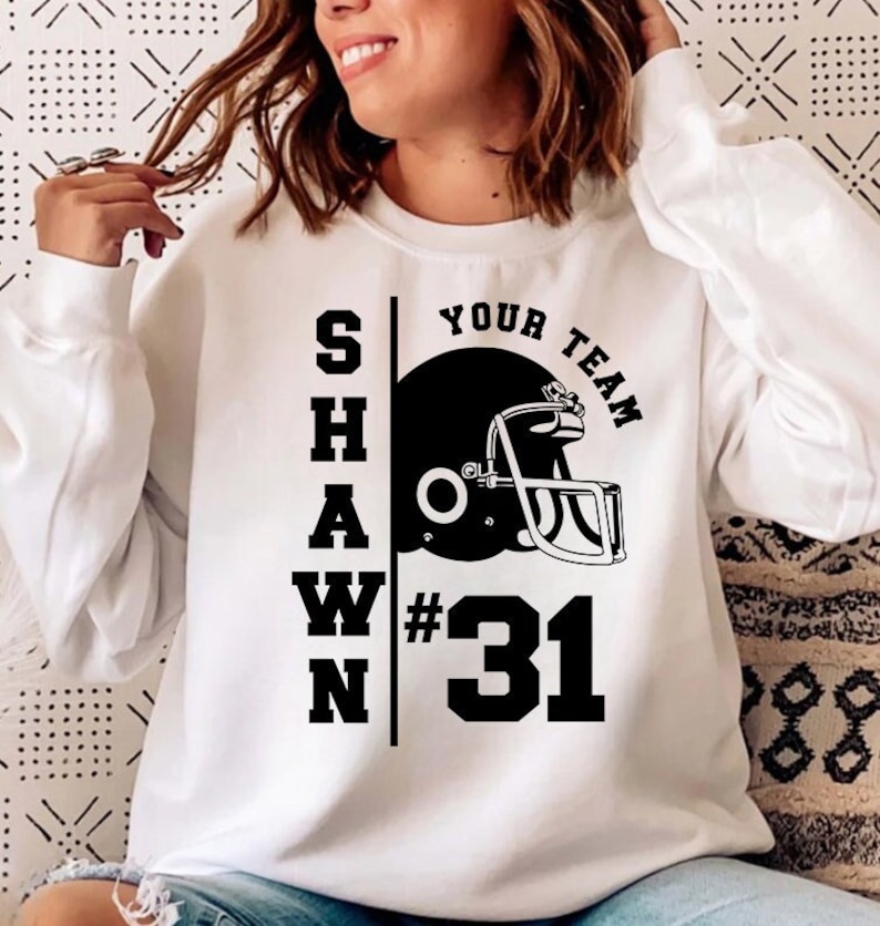 K&ouml;nnte beinhalten: Wei&szlig;er Pullover mit schwarzem Text, der "SHAWN YOUR TEAM #31" lautet, und einer Grafik eines schwarzen und wei&szlig;en Footballhelms.