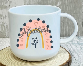 Op de afbeelding: Een witte keramische mok met een regenboogontwerp en de naam "Maddison" in zwarte letters. De regenboog is roze, geel en oranje met een klein groen plantje in het midden. De mok staat op een houten onderzetter.