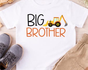 May include: A white t-shirt with the words "BIG BROTHER" in black and orange, featuring a yellow and orange construction vehicle graphic. The shirt is paired with khaki shorts and gray sneakers. Wooden toys are scattered around.