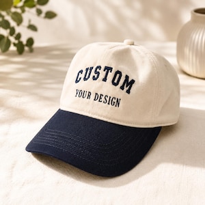 May include: A cream-coloured baseball cap with a navy blue peak. The cap has the embroidered text "CUSTOM" above "YOUR DESIGN" in navy blue. The hat is a classic style, suitable for casual wear.