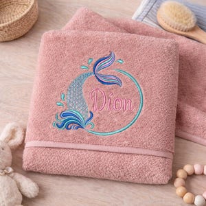 May include: A dusky pink bath towel featuring an embroidered mermaid tail design in blue, teal, and pink. The name "Dion" is embroidered in pink script within a teal circle. The towel is folded with a decorative border.