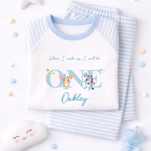 May include: A white and light blue striped pyjama set featuring a graphic of two cartoon dogs with balloons and the text "When I wake up I will be ONE" and the name "Oakley" printed in blue on the front of the long-sleeved shirt.