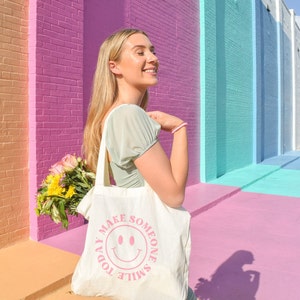 May include: A cream-colored tote bag with the text "TODAY MAKE SOMEONE SMILE" and a pink smiley face. The bag is held by a person wearing a light green dress, standing in front of a colorful brick wall. A bouquet of flowers is visible.