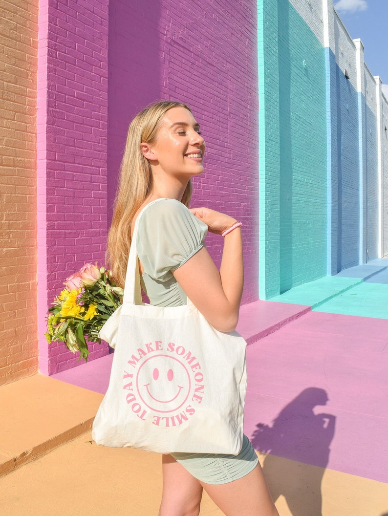 May include: A cream-colored tote bag with the text "TODAY MAKE SOMEONE SMILE" and a pink smiley face. The bag is held by a person wearing a light green dress, standing in front of a colorful brick wall. A bouquet of flowers is visible.