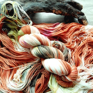 May include: A skein of hand-dyed yarn featuring a variegated color palette of burnt orange, cream, soft sage green, and hints of magenta. The yarn is twisted into a bundle, with loose strands spreading out in a fan-like pattern across the surface.