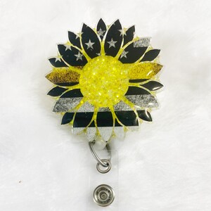 May include: A yellow and black sunflower with a thin yellow stripe across the center. The sunflower is made of resin and has a glitter finish. It is attached to a clear retractable badge holder with silver metal accents.