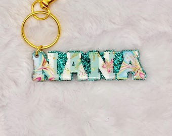 May include: A keychain with the name "NANA" in white block letters. The letters have a floral design with pink and blue accents. The top and bottom of the letters are outlined with teal glitter. The keychain has a gold-colored clasp and ring.