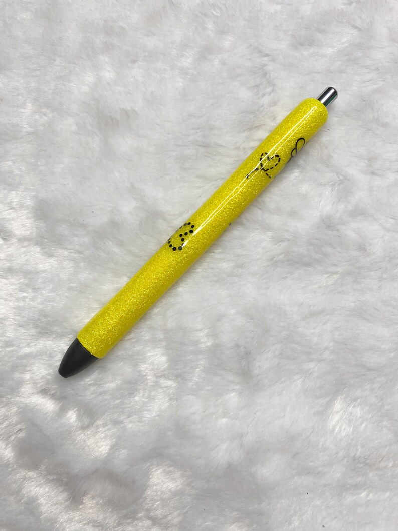 May include: A yellow glitter pen with black ink and a silver clip. The pen has a black smiley face and a black heart design on it.