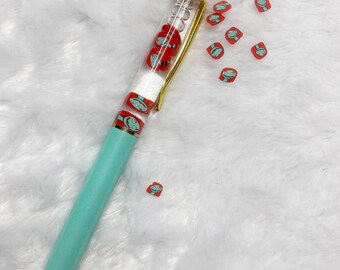 May include: A teal and gold pen with a clear barrel filled with red and white cartoon character figures. The pen has a gold tip and a gold top. Small red and green character stickers are scattered around the pen.
