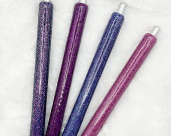 May include: Four glitter pens in shades of purple and blue. The pens have silver tips and black ink tips. The pens are arranged diagonally against a white, fluffy background.