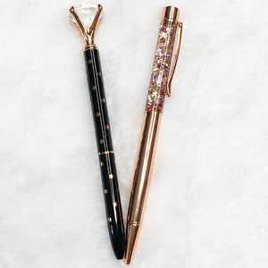 May include: Two elegant pens on a white surface. One pen is black with gold dots and a diamond-shaped top. The other is rose gold with a clear barrel filled with glitter. Both pens have a sleek, modern design.