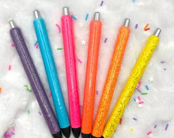 May include: Six colorful pens with glitter barrels in purple, blue, pink, orange, and yellow. Each pen has a silver tip and a black ink cartridge. The pens are arranged on a white, fluffy surface with colorful confetti.