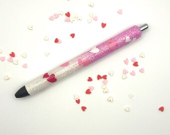 May include: A glittery pen with a pink and white gradient design, adorned with heart-shaped accents. The pen features a silver tip and a black writing end. Scattered around the pen are small heart-shaped decorations in red, pink, and white.