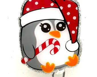 May include: A silver glitter badge reel with a cartoon penguin wearing a red and white Santa hat and holding a candy cane.