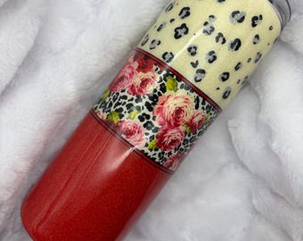 May include: A glittery tumbler with a three-section design. The top section is cream-colored with black leopard spots. The middle section features a floral pattern with red roses. The bottom section is red.