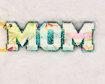 May include: A decorative keychain spelling out "MOM" in capital letters. The letters are filled with a floral pattern in shades of pink, blue, and green. The edges of the letters are outlined with teal glitter, and a gold-colored keyring is attached.