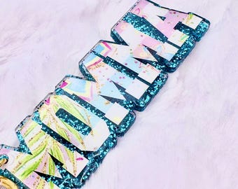 May include: A set of resin keychains spelling out the word "HOME". Each letter is a different color and pattern, with a teal glitter border. The letters have a floral and pastel design.