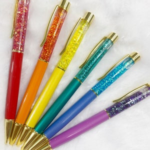 May include: Six glitter pens with gold accents and clear barrels filled with colorful glitter. The pens are arranged in a rainbow pattern, with red, orange, yellow, green, blue, and purple.