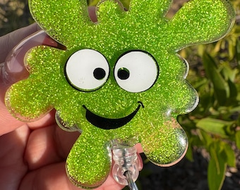 May include: A green glitter badge reel in the shape of a smiling blob with white eyes and a black smile. The badge reel has a clear plastic retractable cord and a metal clip.