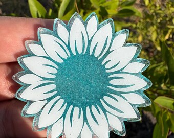 May include: A sunflower-shaped resin charm with a white petal design and a teal glitter center. The charm has a clear, slightly iridescent border. The background is blurred, with green foliage.