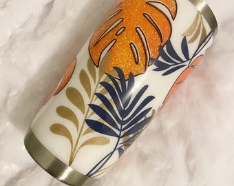 May include: A white tumbler with a stainless steel rim and a tropical leaf design. The design features orange glitter monstera leaves, gold and navy blue foliage, and coral accents.