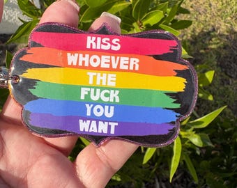 May include: A colorful keychain with a rainbow design and the text "KISS WHOEVER THE FUCK YOU WANT". The keychain has a glittery border and a metal ring for attachment. The background is blurred green foliage.