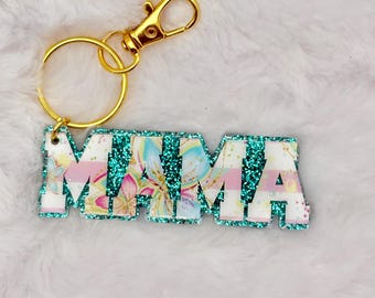 May include: A keychain with the word "MAMA" in white block letters. The letters are decorated with a floral pattern and teal glitter. The keychain has a gold-colored clasp and ring.