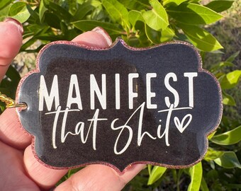 May include: A black and glittery keychain with the words "MANIFEST That Shit" in white script. The keychain has a decorative shape and a gold-colored loop for attachment. The background is blurred green foliage.