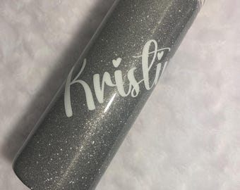 May include: A silver glitter tumbler with a clear lid. The name "Kristi" is written in white script on the side of the tumbler. The tumbler is cylindrical and appears to be made of stainless steel.