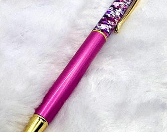 May include: A pink pen with gold accents. The pen's barrel is a vibrant pink, while the tip and the top are gold. The upper section of the pen is filled with a clear liquid and colorful glitter. The pen has a gold clip.