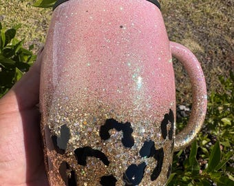 May include: A pink glitter mug with a black lid and handle. The bottom half of the mug transitions to gold glitter with black leopard print spots. The mug is designed for beverages.