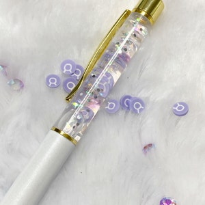 May include: A white pen with a clear barrel filled with iridescent glitter and purple confetti. The pen has a gold top and a gold band around the barrel.
