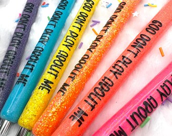 May include: A set of six glittery pens in purple, blue, yellow, orange, and pink, each featuring the black text GOD DON'T PLAY ABOUT ME. The pens have black tips and silver-toned metal accents at the base, arranged in a diagonal row.