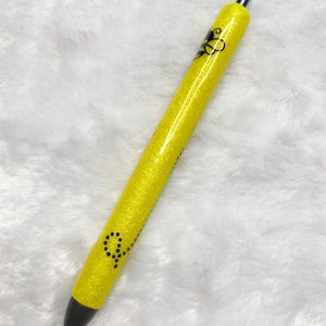 May include: A yellow glitter pen with a black bee design and black dots. The pen has a silver clip.