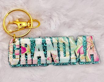 May include: A keychain with the word "GRANDMA" in white block letters. The letters are set against a floral pattern with pink and green accents, and outlined with teal glitter. The keychain hardware is gold.