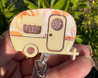 May include: A badge reel featuring a camper design. The camper is cream-colored with a floral pattern on the roof and glitter accents. The badge reel has a clear plastic strap and a metal clip for attaching to clothing.