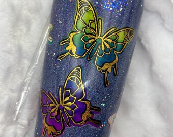 May include: A glittery blue tumbler with two butterfly designs. The butterflies have gold outlines and gradient wings in shades of green, blue, and purple. The tumbler has a black lid and is covered in sparkling glitter.