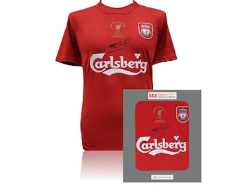 Może przedstawiać: Czerwona koszulka piłkarska Liverpoolu z logo Carlsberga i herbem klubu. Koszulka jest podpisana przez zawodnika i ma złotą naszywkę upamiętniającą zwycięstwo klubu w Lidze Mistrzów. Koszulka jest prezentowana w oprawie.