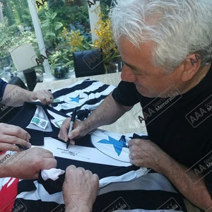 May include: A black and white soccer jersey with a blue star on the front is being signed by a person. The jersey is laid out on a table and the person is holding a black pen.