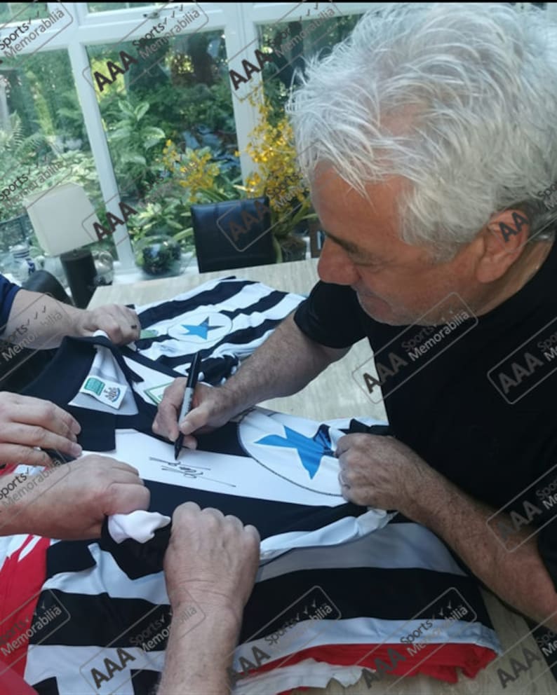 May include: A black and white soccer jersey with a blue star on the front is being signed by a person. The jersey is laid out on a table and the person is holding a black pen.