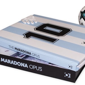 Op de afbeelding: Twee hardcover boeken getiteld "The Maradona Opus" zijn op elkaar gestapeld. Het bovenste boek heeft een lichtblauw en wit gestreept ontwerp met het nummer 10. Een voetbal met zwarte en blauwgroene accenten ligt naast de boeken. De boeken staan op een witte achtergrond.