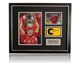 May include: Framed sports memorabilia featuring a player in a red jersey holding a trophy. The frame includes two photos, a signed yellow card, and a plaque that reads "Personally Signed by STEVEN GERRARD Liverpool FC Captain."