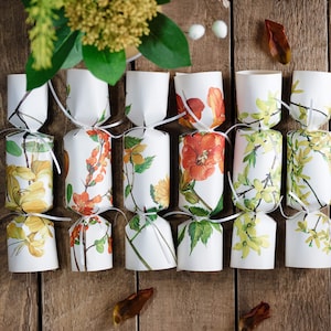 May include: Eight white paper Christmas crackers with floral designs. The crackers have a yellow, orange, and green floral pattern. The crackers are tied with white ribbon.