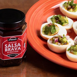 May include: A plate of deviled eggs topped with guacamole, red onion, and cilantro. A jar of "Valente Artesanal Hot Sauce Salsa Brava De Chiles Secos" is in the foreground.