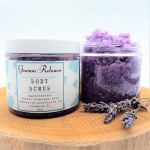 Op de afbeelding: Een paarse bodyscrub in een glazen pot met een wit deksel. De pot is gelabeld "Gewoon Relaxen BODY SCRUB" en vermeldt de ingrediënten: "Suiker. Himalaya zout Kokosolie.Castorolie en Vitamine E." De pot staat op een houten oppervlak met een takje lavendel.